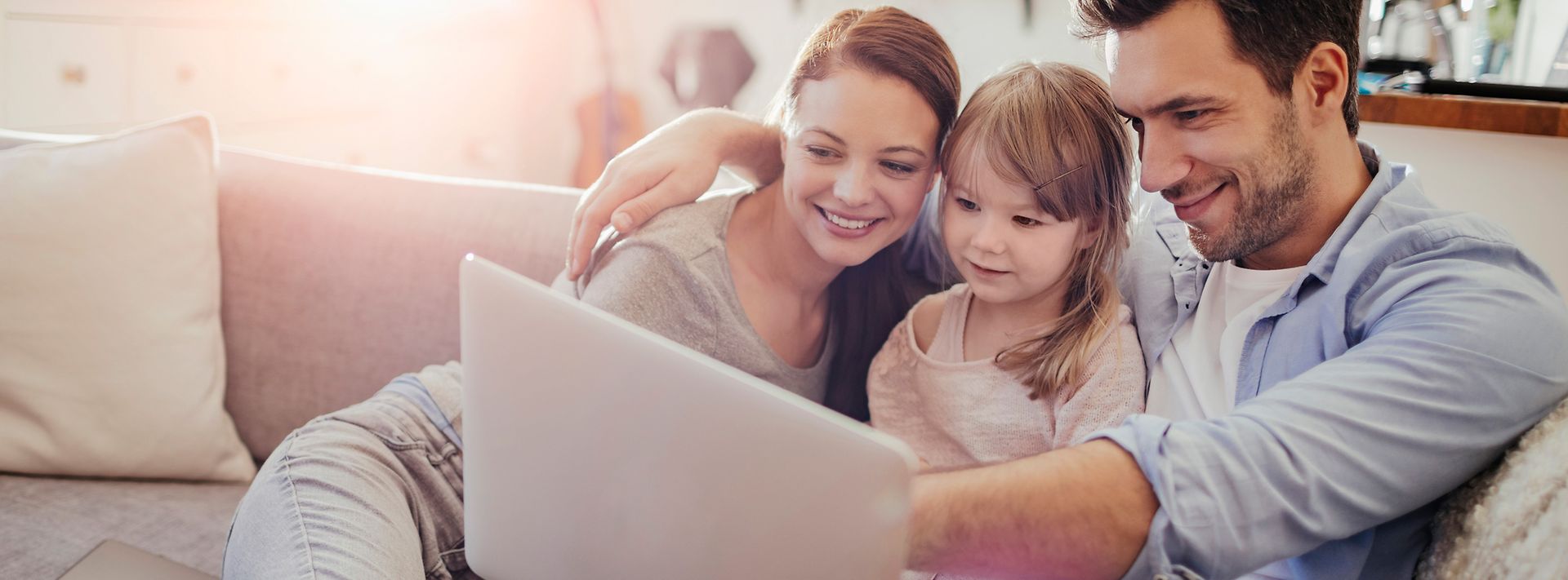 © iStock.com/Geber86 Frau und Mann mit kleiner Tochter sitzen eng zusammen auf dem Sofa und schauen auf einen Laptop