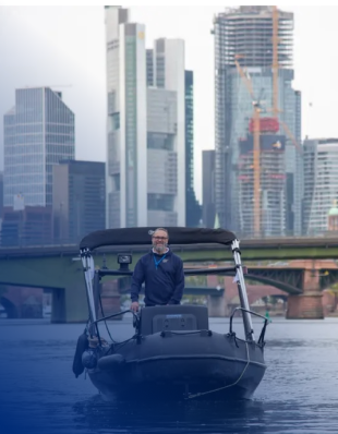 Mann steht am Steuer eines Sportboots im Wasser, im Hintergrund Skyline einer Stadt