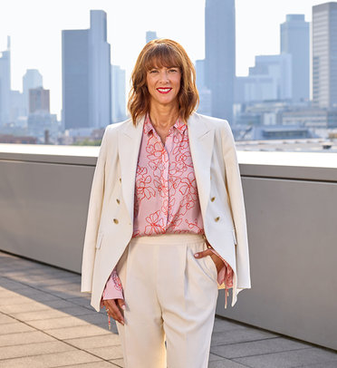 Frau in weißem Blazer mit rosa Bluse und Muster, stehend vor Skyline. Freundlicher Gesichtsausdruck.