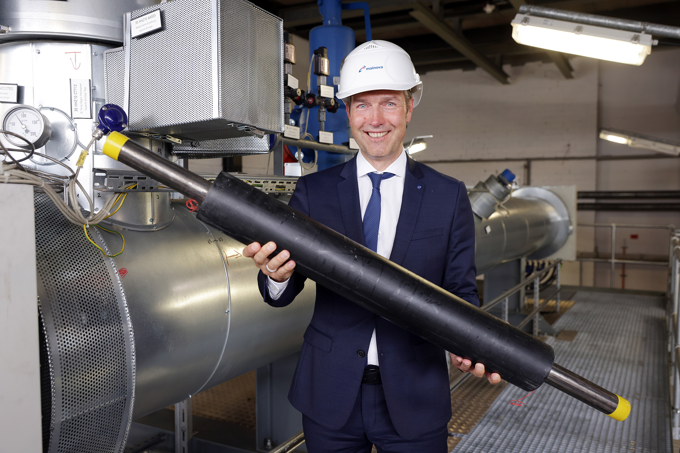 Mainova-Vorstand Martin Giehl trägt Helm und hält eine große, zylindrische Metallröhre in einer industriellen Umgebung.