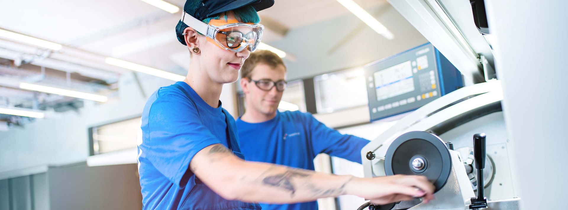 Junge Frau steht an Drehmaschine und der Ausbilder schaut der Arbeit zu.