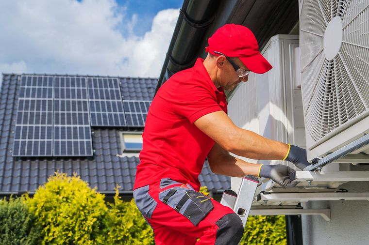 Mann installiert Wärmepumpe im Hintergrund ist ein Hausdach mit PV-Anlage