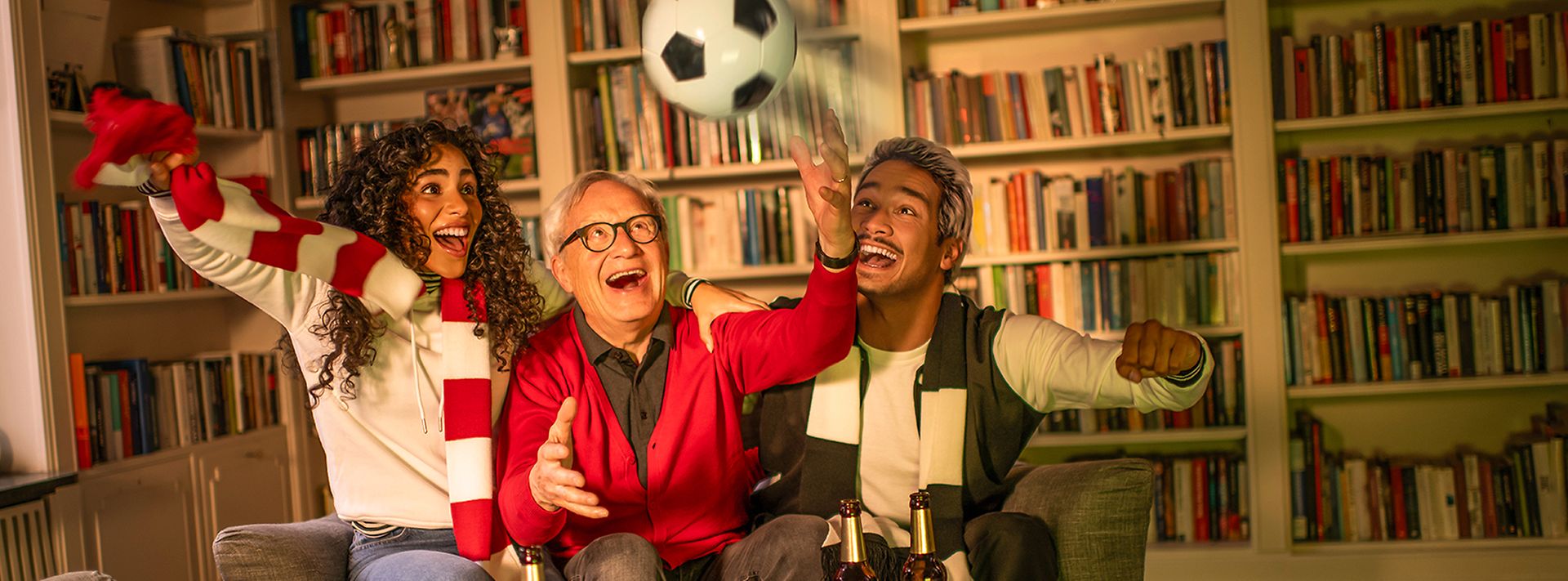 Drei Menschen jubeln zusammen auf einem Sofa, während sie ein Fußballspiel schauen. Ein Fußball fliegt durch die Luft.