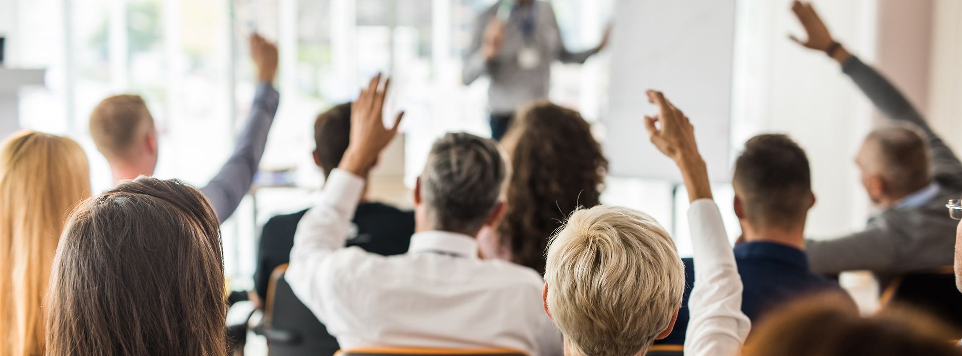 Personengruppe in einem Raum versammelt, stellt Fragen an eine Person die mit einem Flipchart eine Präsentation hält 