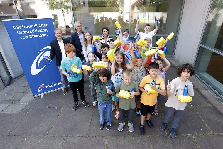 Eine Gruppe von Kindern mit gelben Brotboxen, vier Erwachsene und ein Mainova Aufsteller im Hintergrund