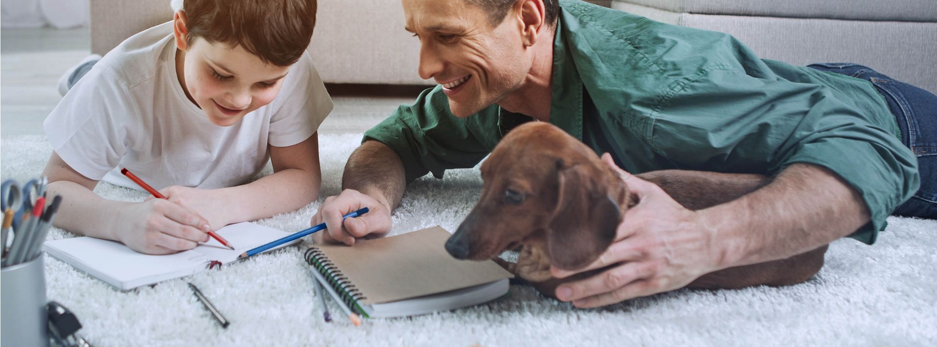 Vater, Sohn und Dackel liegen auf dem Wohnzimmerteppich und machen schriftliche Hausaufgaben