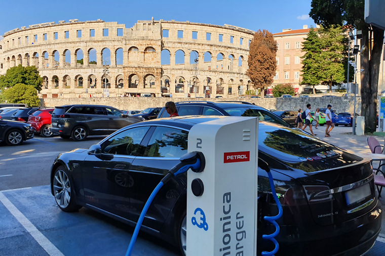 E-Auto steht an einer Ladesäule, ein Amphitheater im Hintergrund.